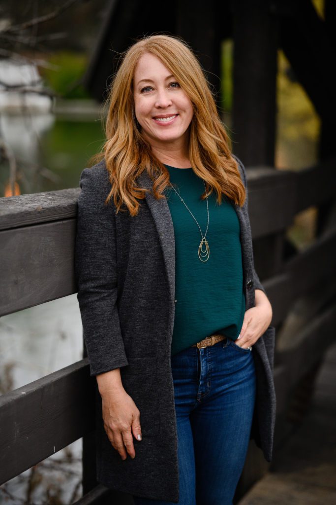 Denver Commercial Photographers capture a red headed woman in a dark gray blazer and green shirt leaning against a bridge with her hand in her pocket.