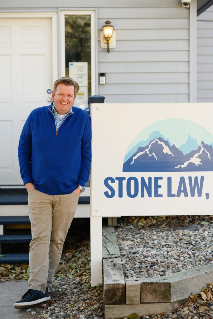 Man in bright blue sweater leaning against a white sign that says Stone Law for Denver Commercial Photographers.
