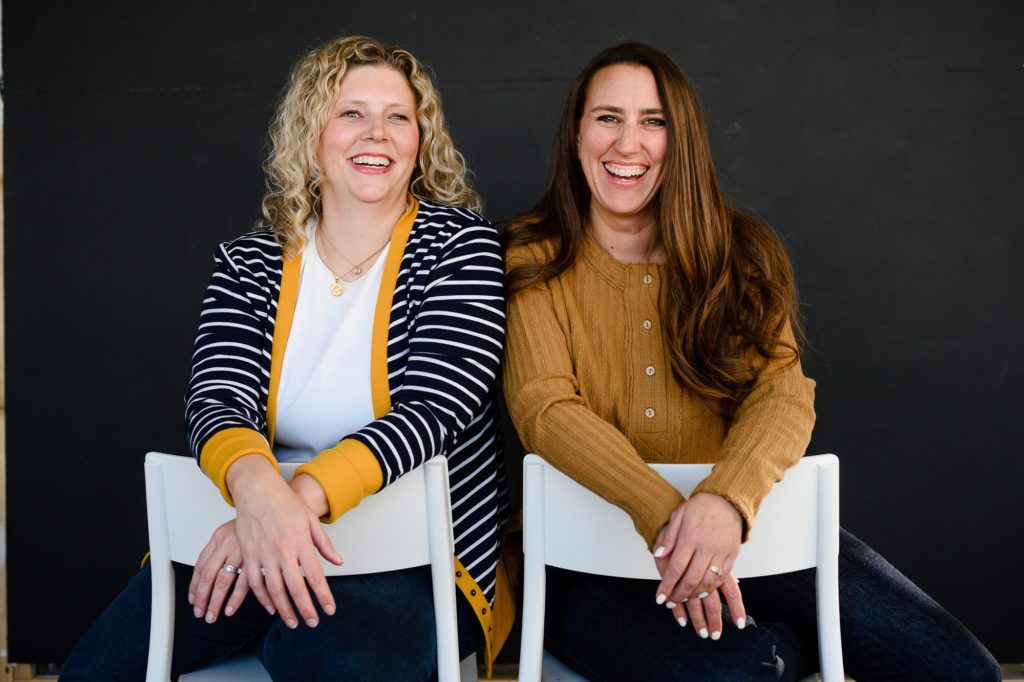 A blonde woman and a brunette woman sit backwards in white chairs smiling at Denver Commercial Photographers