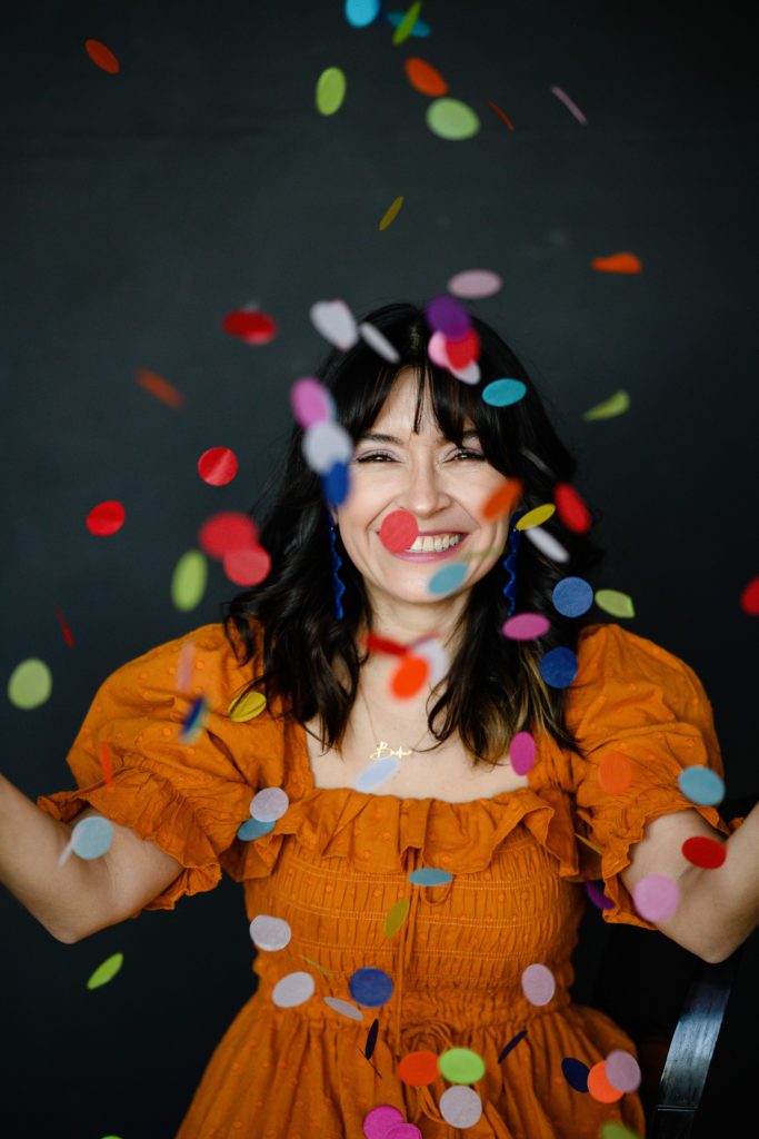 Denver Commercial Photographers capture a latina woman in an orange dress throwing brightly colored confetti dots in the air and smiling.
