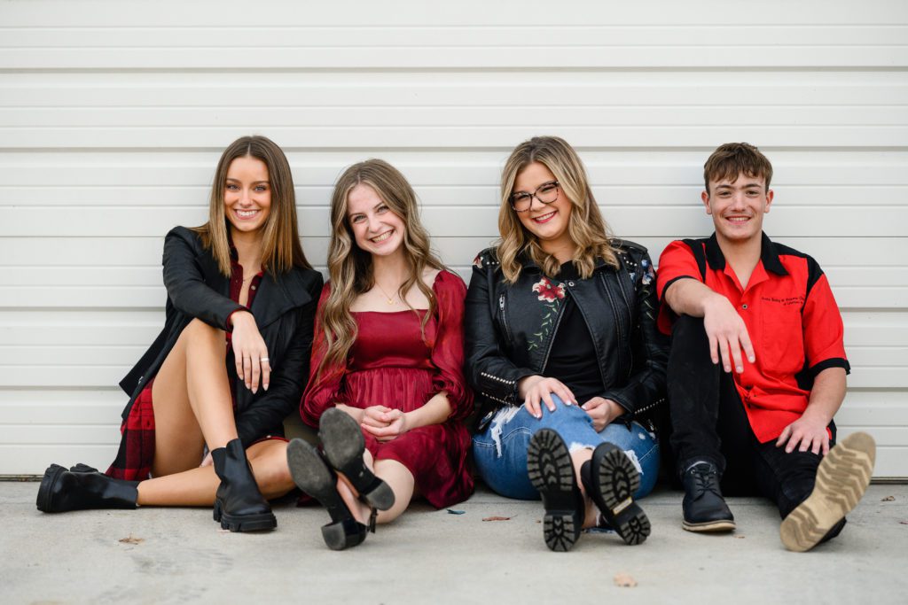 A group of 4 teens sitting in front of a white garage door smiling at a denver senior photographer