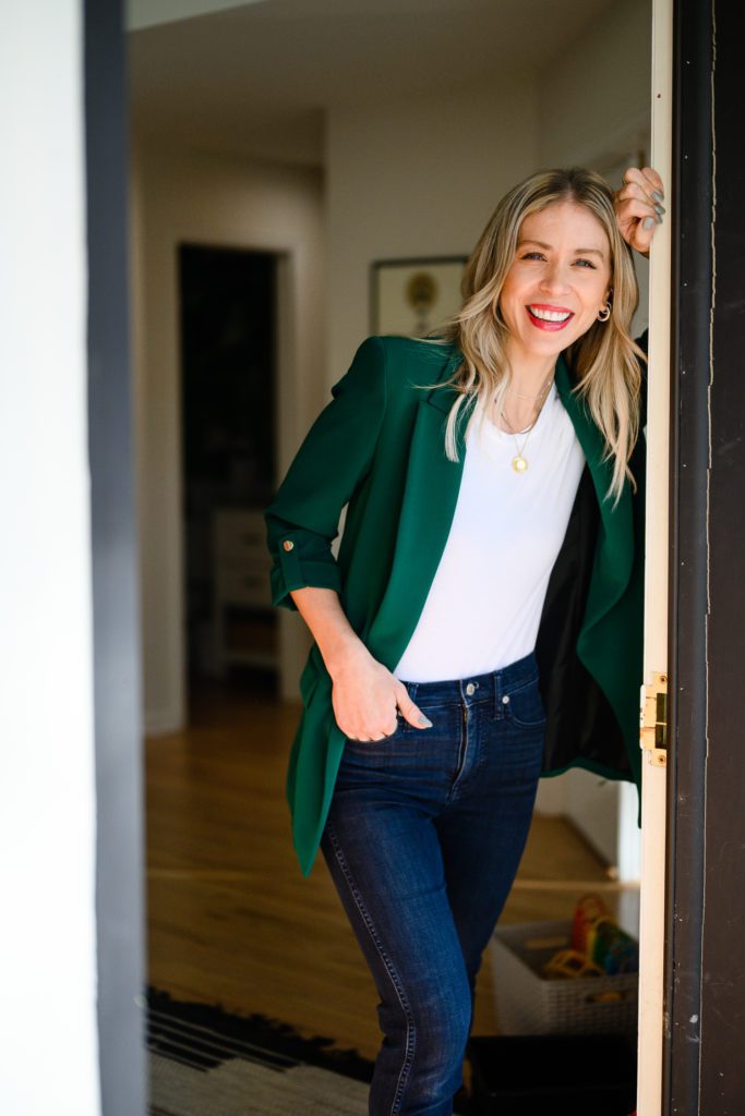 A woman in a green blazer leaning on her front doorway smiling at Denver commercial photographers for her branding photos.