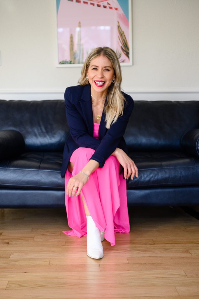 A woman in a bright pink dress and blue blazer sitting on a couch smiling at Denver commercial photographers for her branding photos.