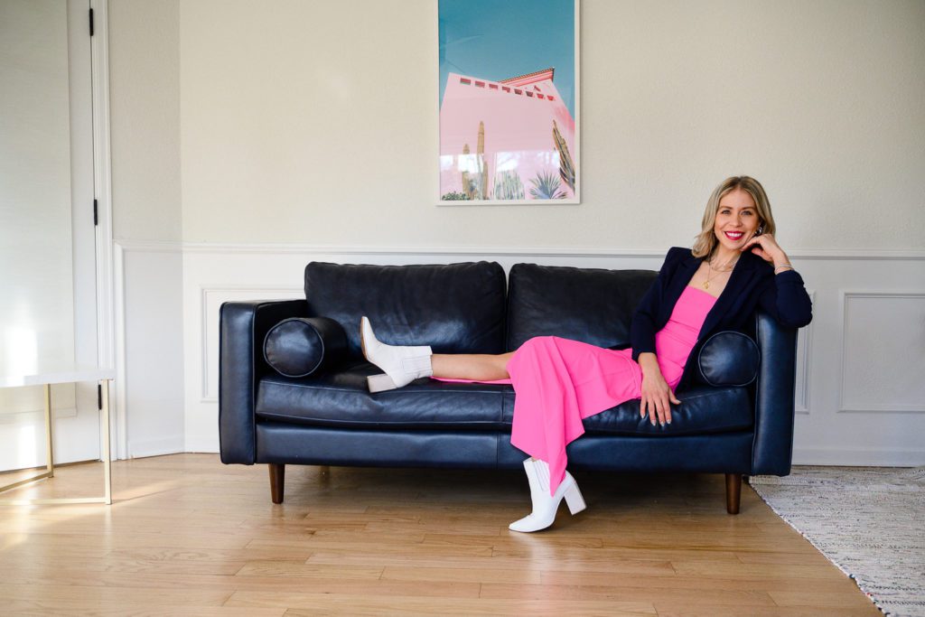 A woman in a bright pink dress lays on couch with blue blazer and white boots smiling at Denver commercial photographers.