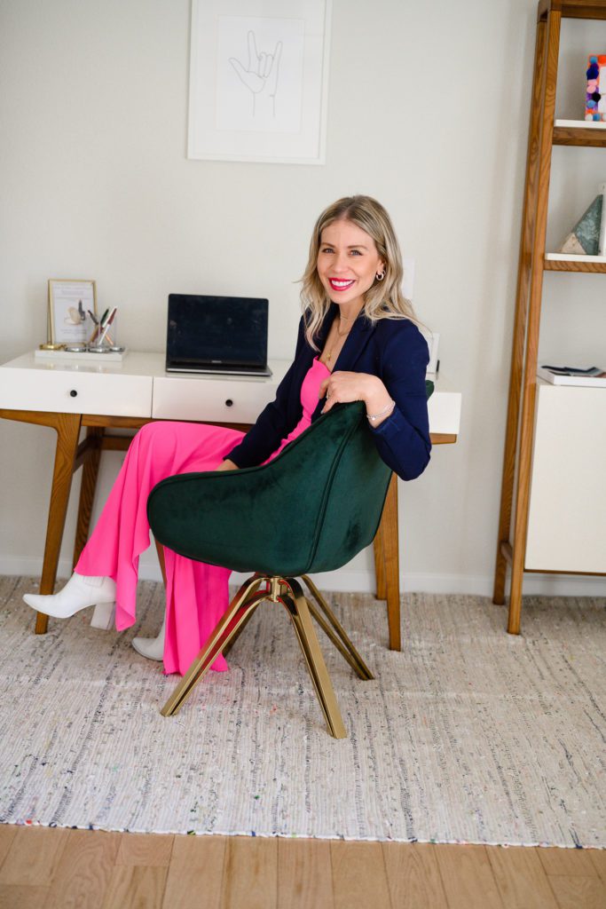 Denver Branding Photographer captures a woman in a bright pink dress and blue blazer sitting at her desk in her studio office.