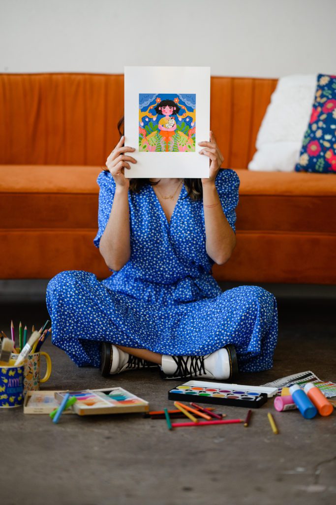 Denver Commercial Photographers captures woman covering her face with art wearing a vibrant blue dress with art supplies in front of her.