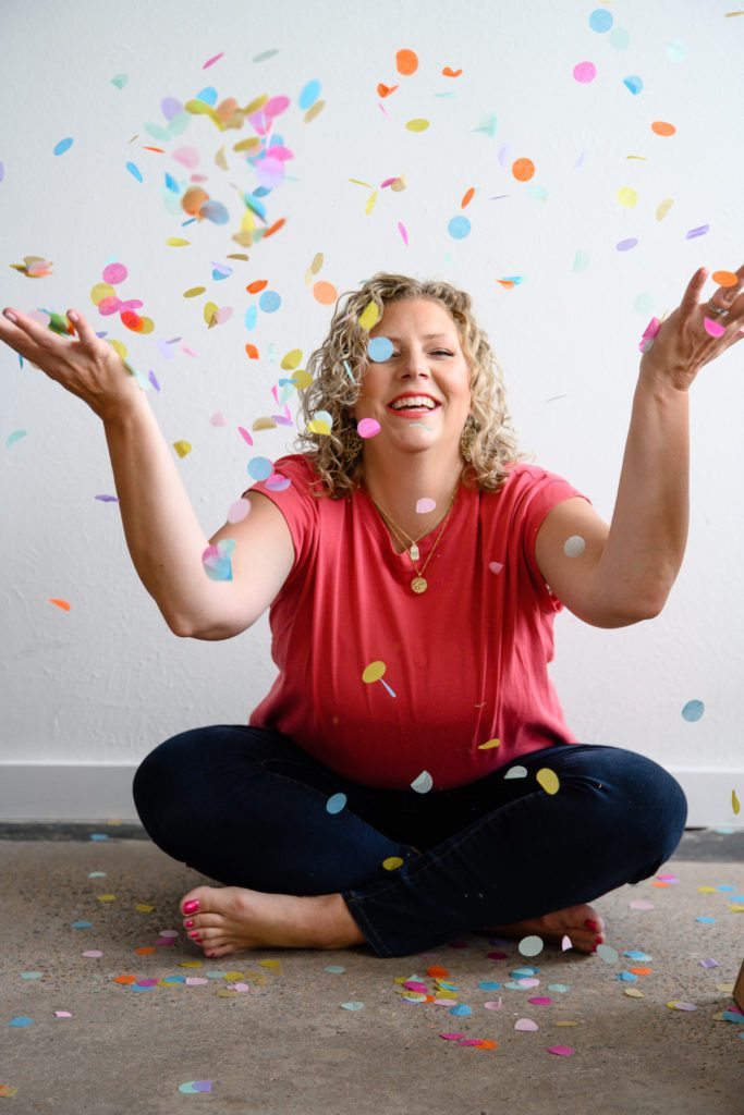 Denver Commercial Photographers captures woman smiling at camera and throwing confetti in the air wearing a pink shirt and jeans.