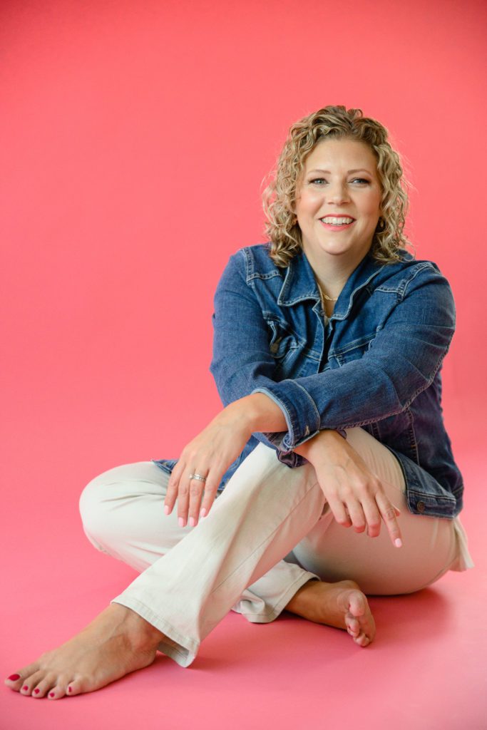 Denver commercial photographers capture a woman with a bright pink backdrop sitting crosslegged wearing a jean jacket and khaki pants.