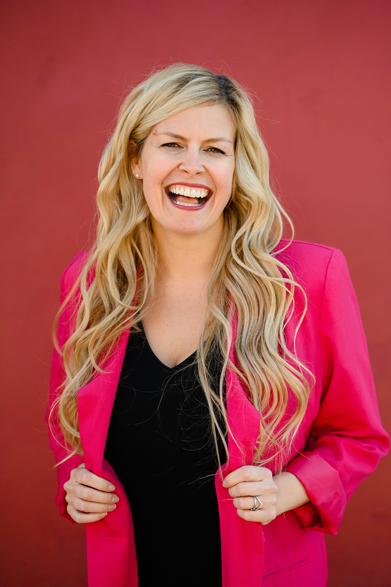 prepare for branding photos with denver commercial photographer with woman in a a hot pink blazer laughing as she stands against a red background for her headshots