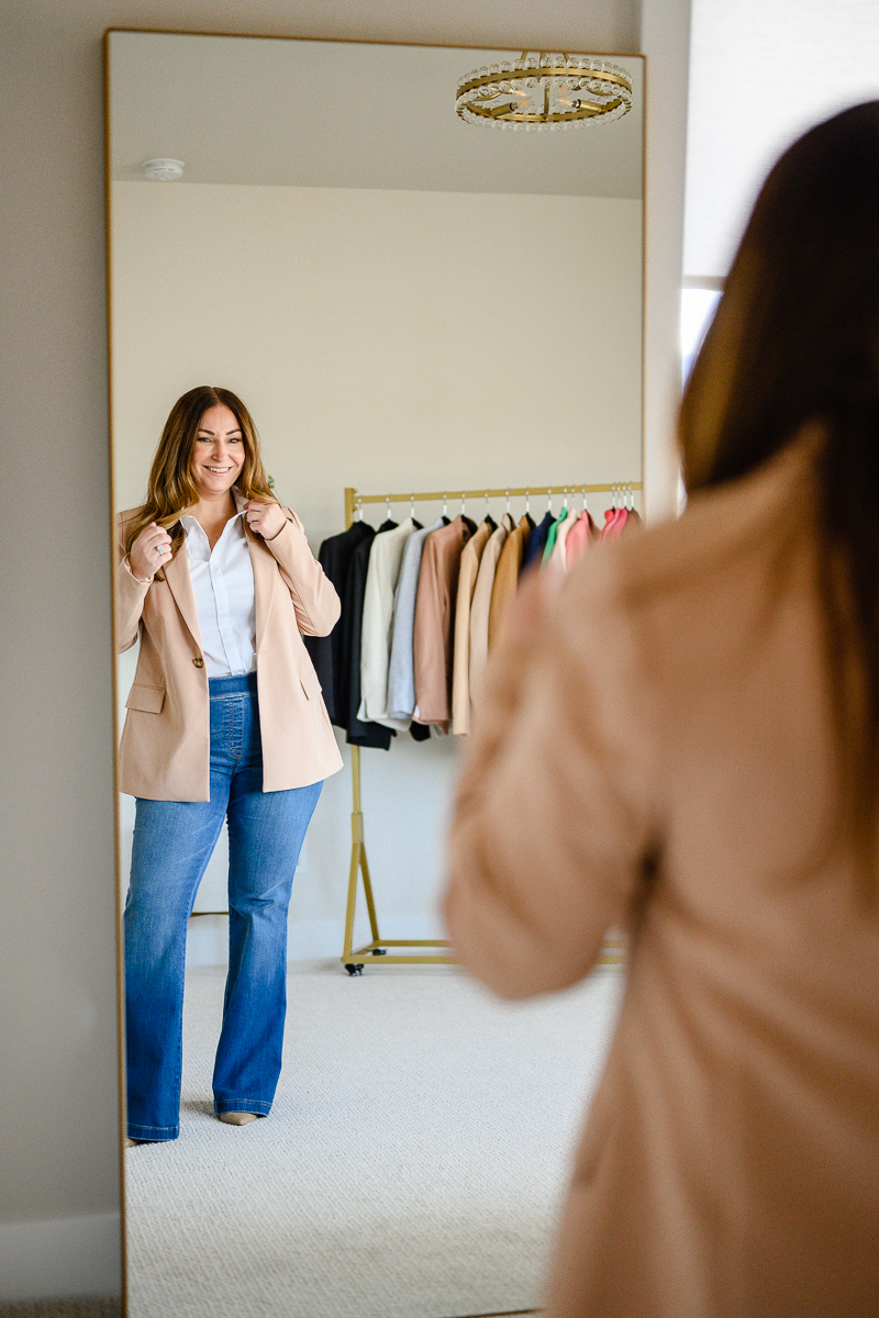 Brand pictures by Denver commercial photographers with woman putting on a suit coat while standing in the mirror and posing with a rack of coats behind her