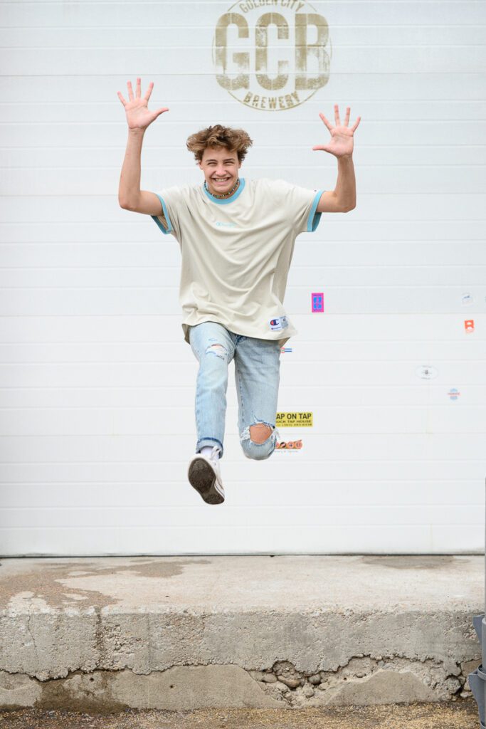 denver senior photographer captures fun senior pictures with young man jumping off of a concrete ledge with his hands in the air