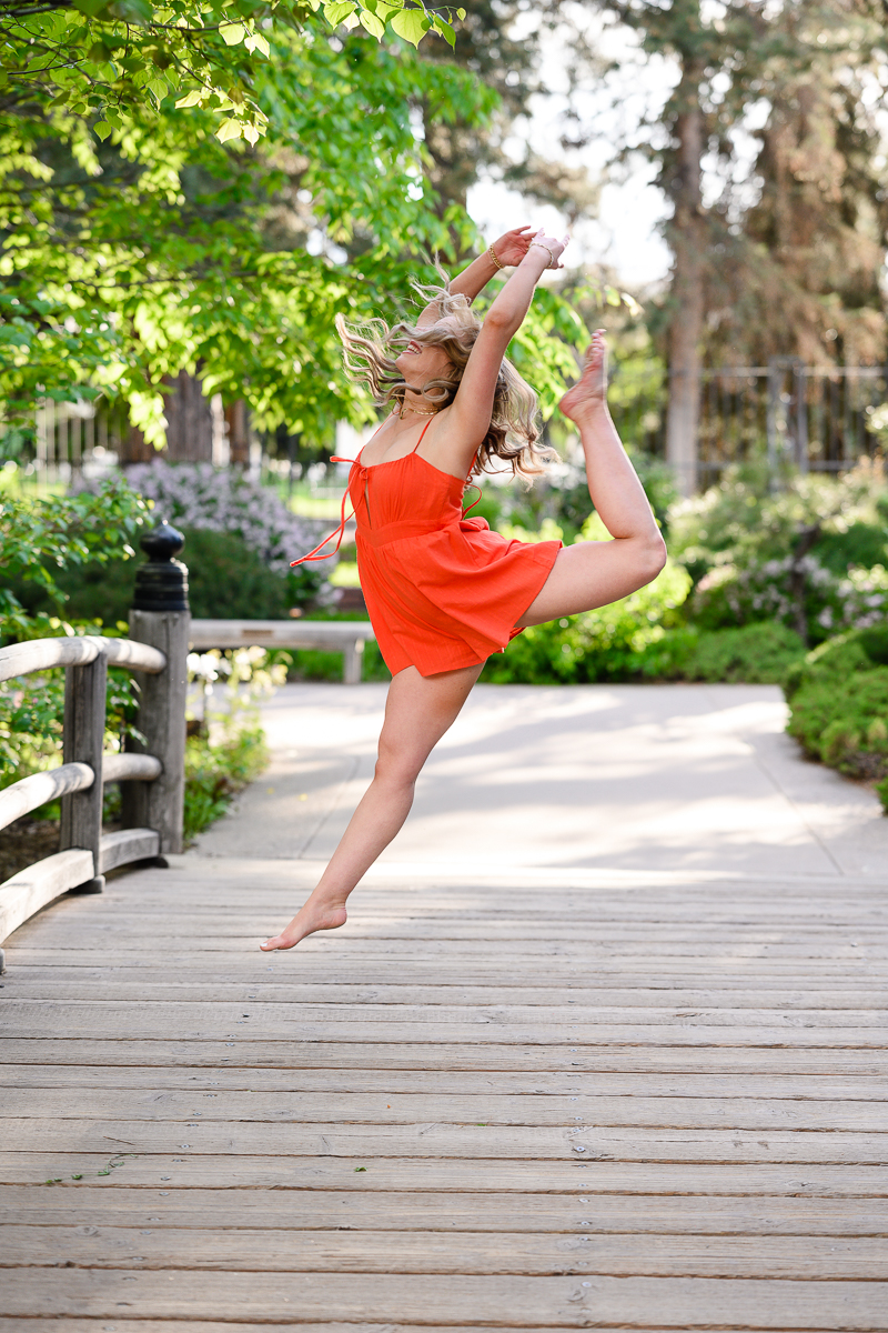 creative senior pictures with girl in a bright orange dress dancing on a bridge with a lush garden behind her