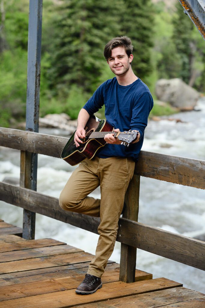 A senior boy leans on a bridge in Aspen that overlooks the river and plays his guitar for a Denver senior photographer