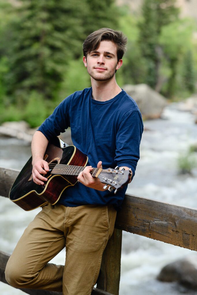Denver Senior photographer captures A senior boy leans on a bridge in Aspen that overlooks the river and plays his guitar
