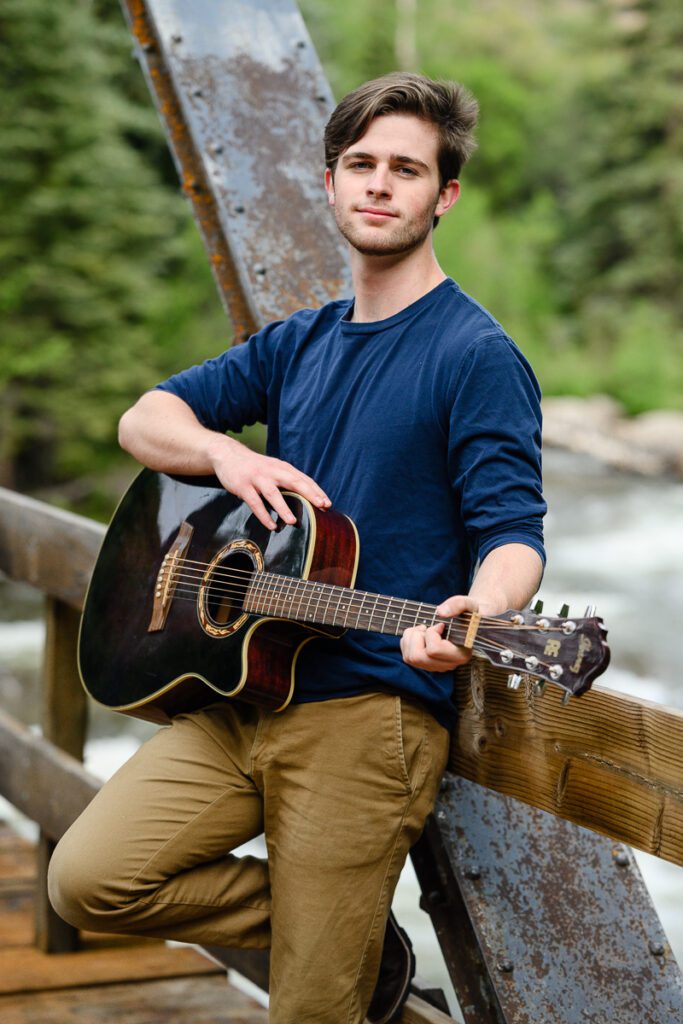 A river in Aspen with a bridge over it holds a young man playing his guitar for his Denver Senior Photographer