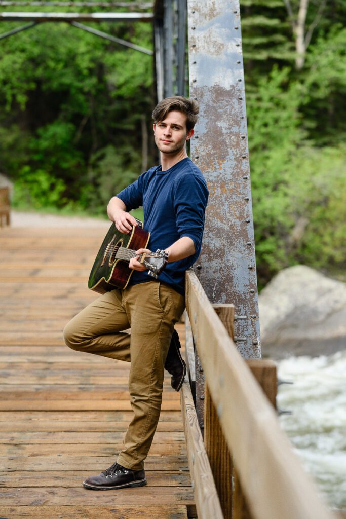 A wide shot of a senior guy standing on a bridge and holding a guitar in Aspen Colorado for his Colorado Senior Pictures.