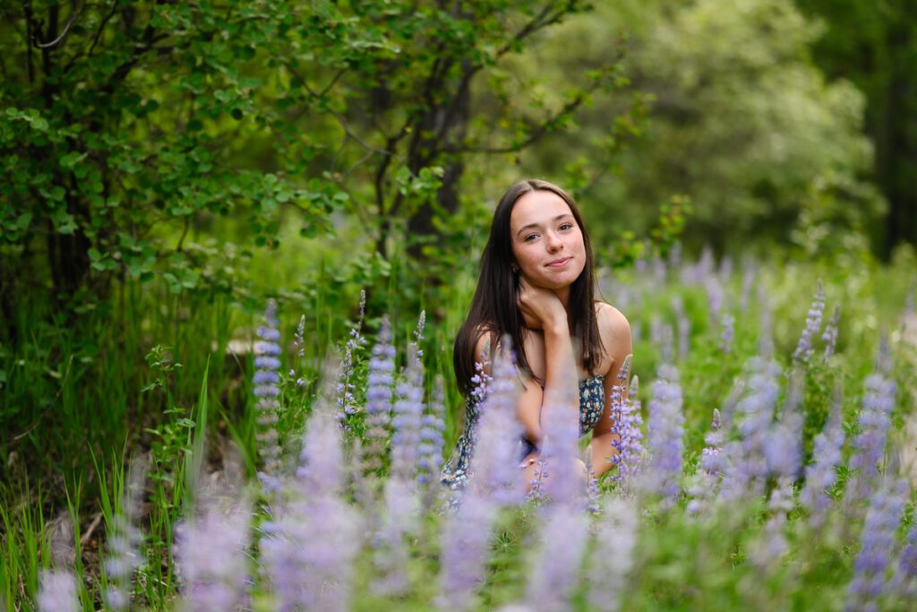 A Denver Senior girl smirks at the camera of a Denver Colorado senior photographer while sitting in a field of purple wildflowers