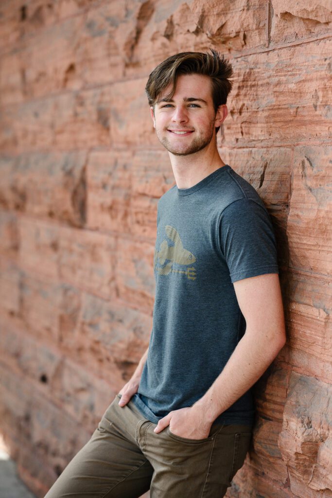 A senior guy standing in front of a red stone wall in Aspen Colorado for his Colorado Mountain senior pictures taken by a Denver senior photographer