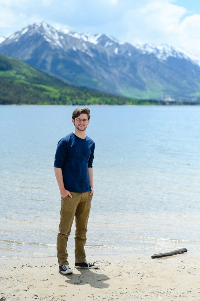 A senior guy stands in front of twin lakes outside of Aspen Colorado for his Colorado mountain senior pictures as taken by Denver senior photographer