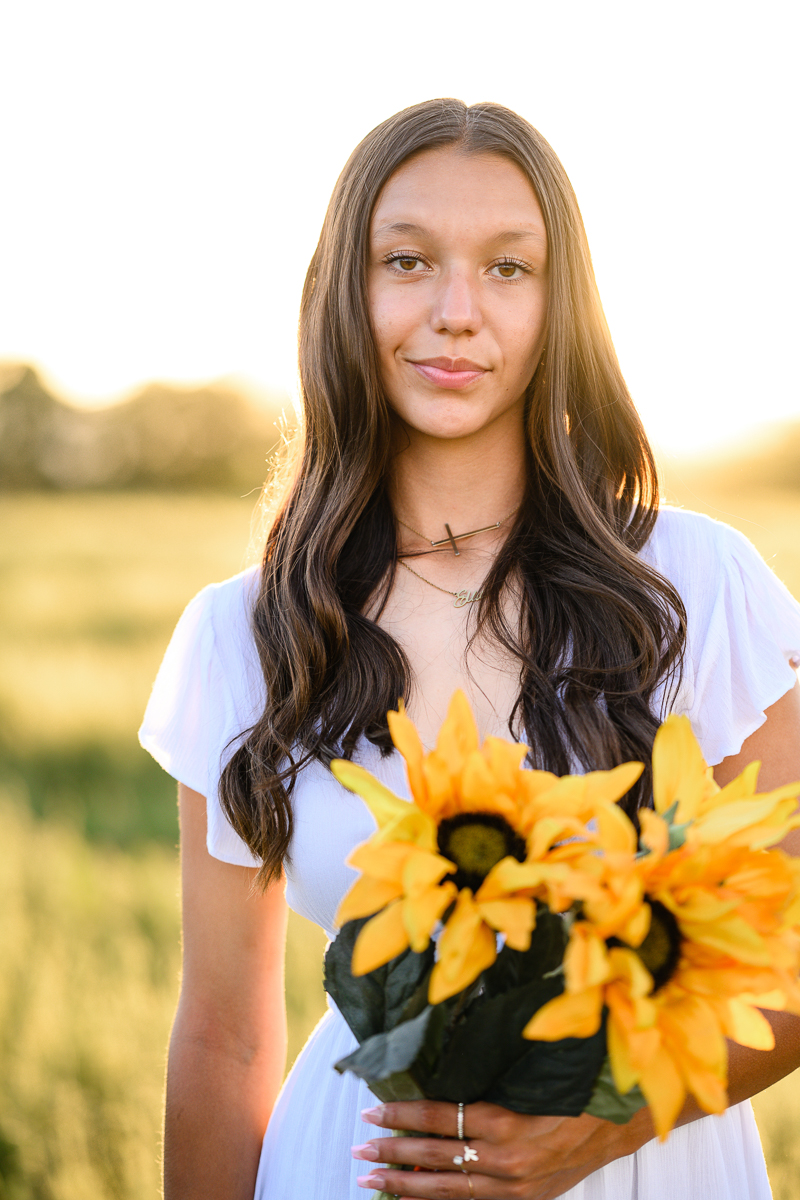 sunset senior session with brunette girl holding sunflowers in a field while in a white dress for her senior picture outfits captured by denver senior photographer