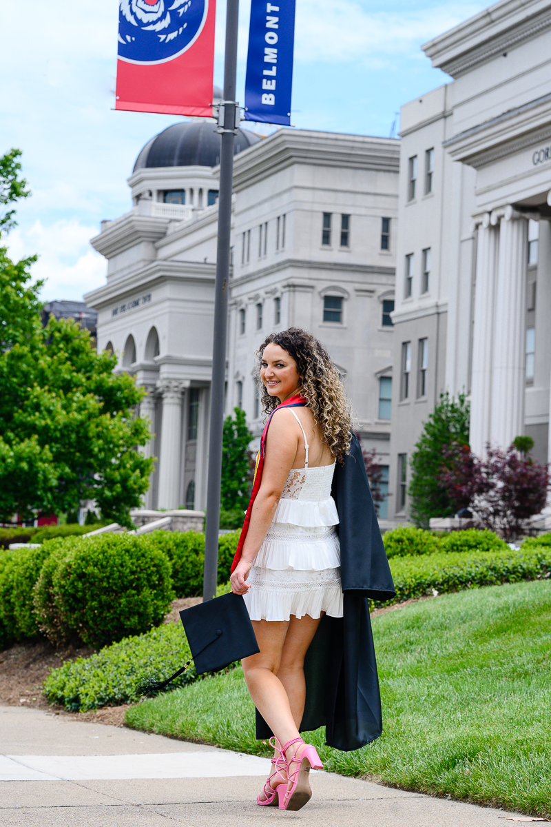 graduation pictures with woman holding her cap and gown and walking onto a campus while looking over her shoulder and smiling captured by denver senior photographer