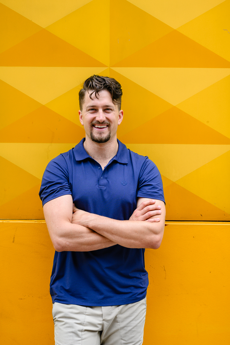 branding photography by Denver commercial photographer with man in a navy blue shirt with his arms crossed standing against a bright yellow wall for his headshots