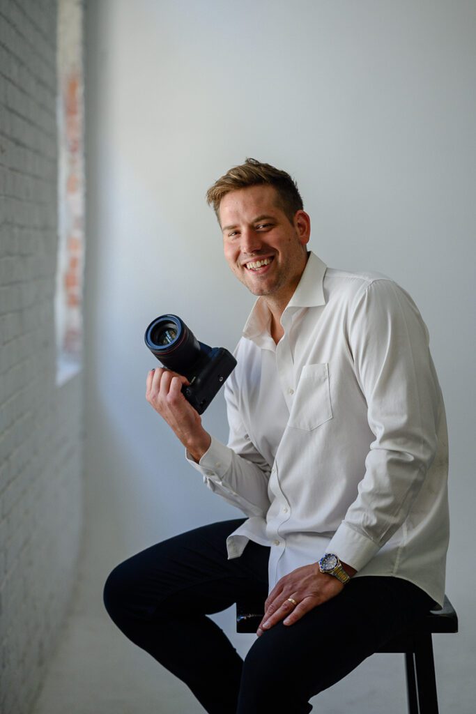 A man holding a camera sitting on a stool smiles at a Denver Commercial Photographer.