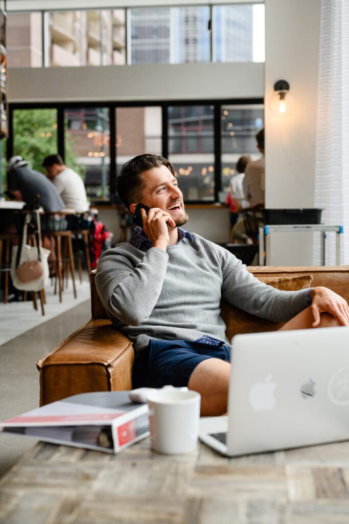 A man sits in a leather chair talking on the phone in the middle of a busy coffee shop as captured by a Denver branding photographer.