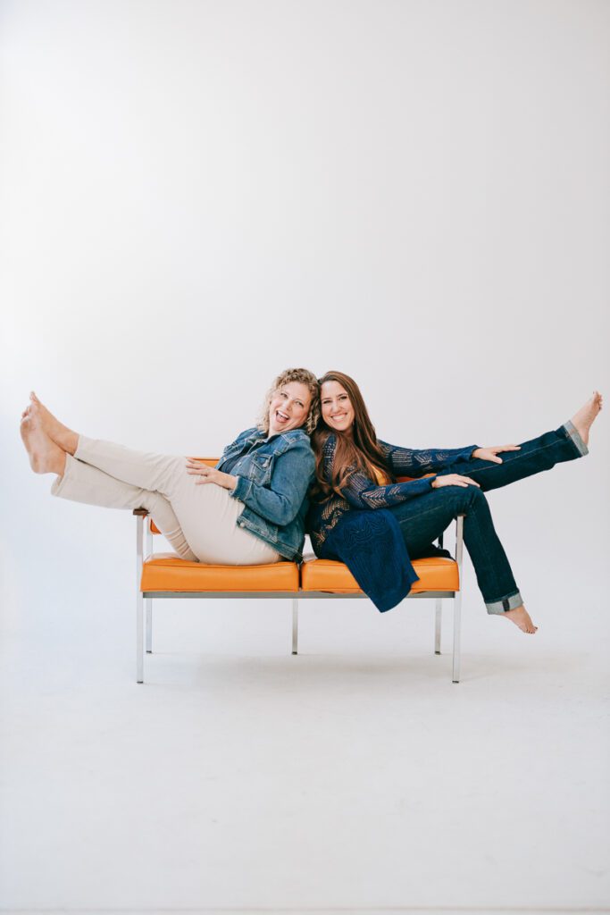 Two women sitting back to back on an orange coach for their Denver Branding photos taken by a Denver Branding photographer and brand strategist.