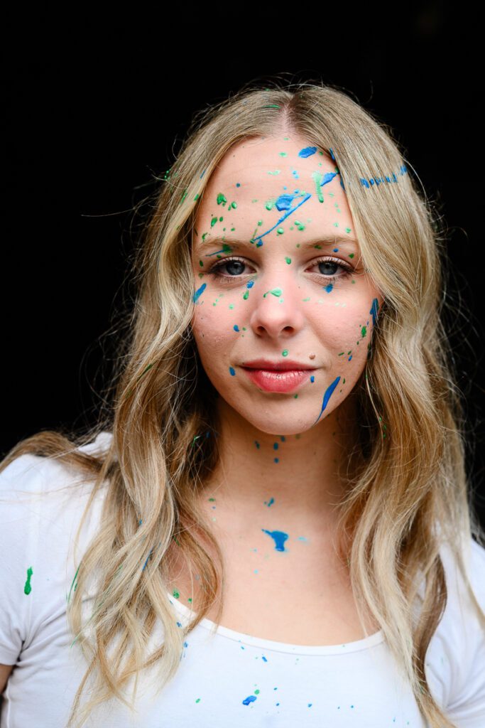 A young woman looks at a Denver photographer while having paint all over her face for her Denver Senior Photos.