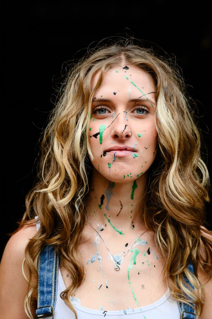 A young woman looks at a Denver photographer while having paint all over her face for her Denver Senior Photos.