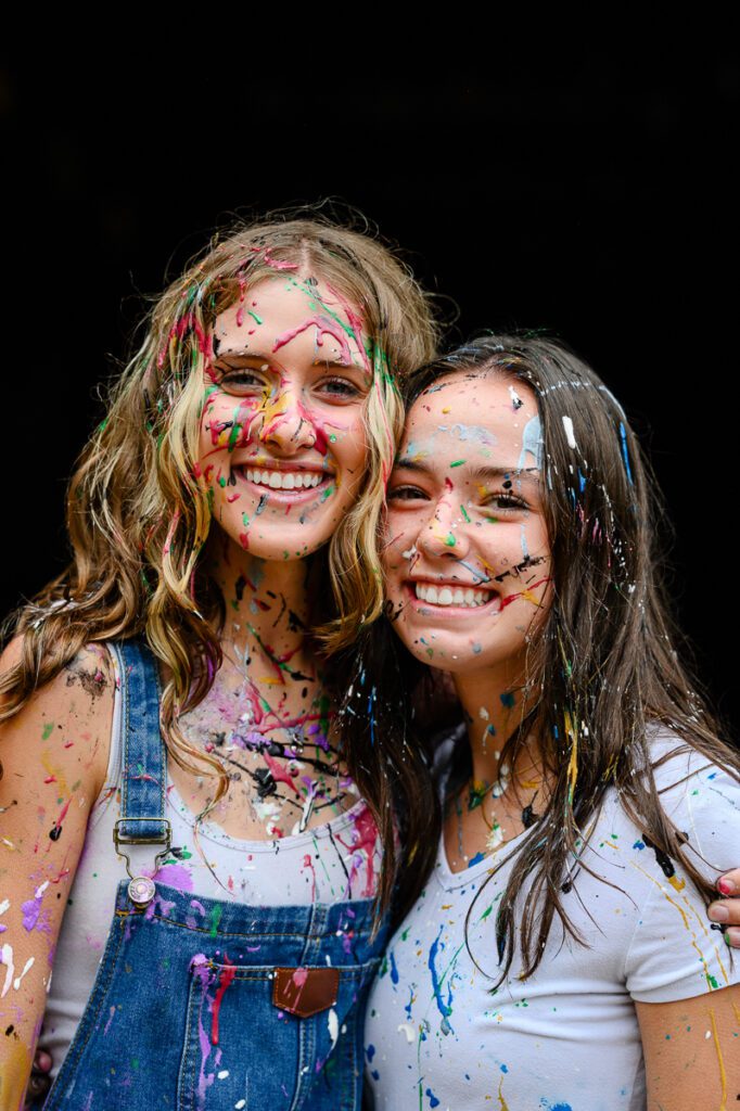 Two young woman look at a Denver photographer while having paint all over her face for her Denver Senior Photos.