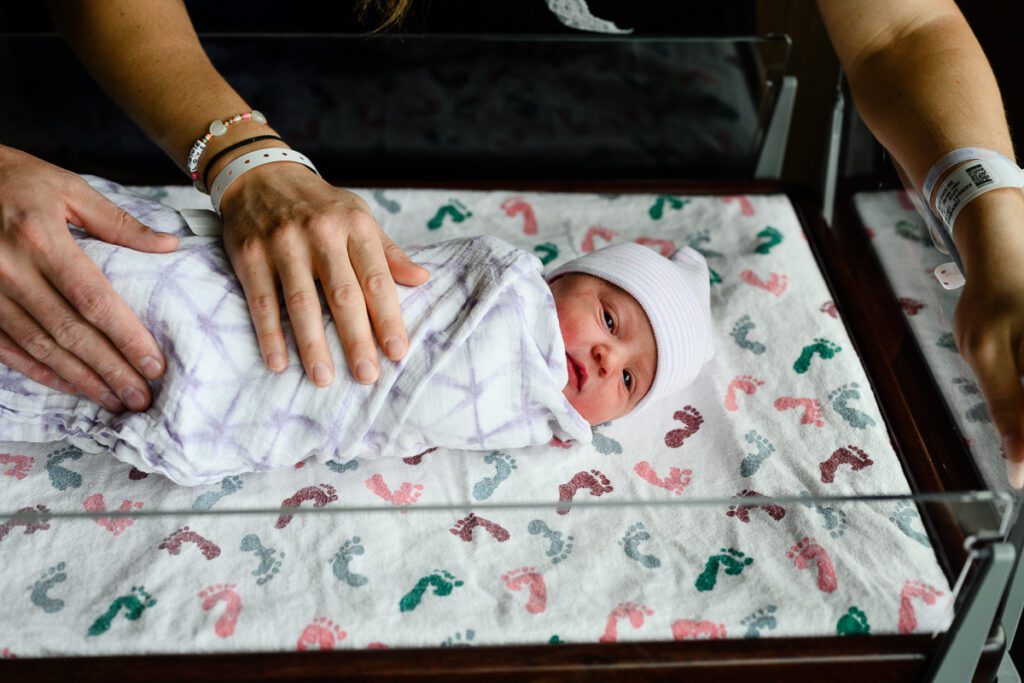 A baby in a hospital bed all wrapped up with her parents hands on her belly captured by a Denver photographer.