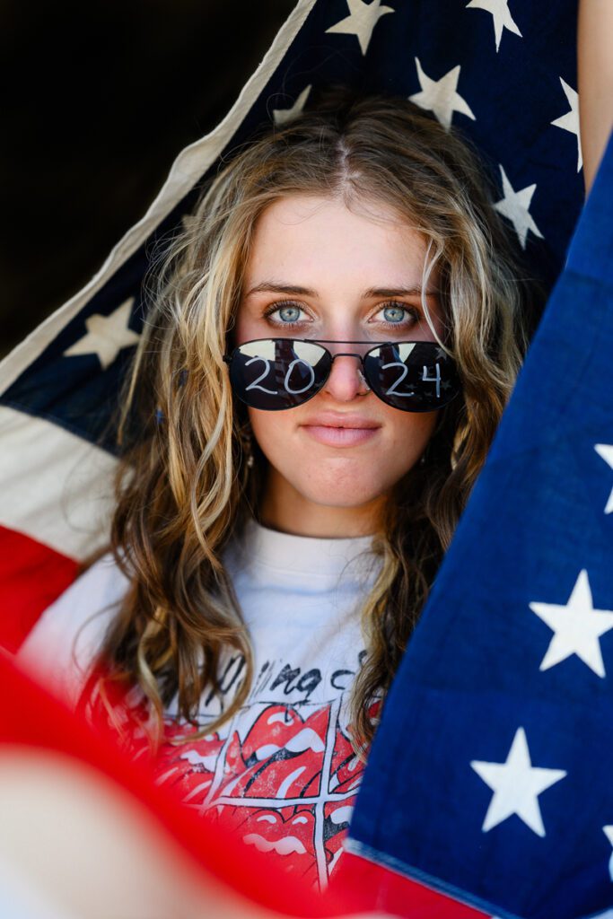 A senior wearing sunglasses that say 2024 holding an american flag as captured by a Denver photographer and Denver Branding expert.