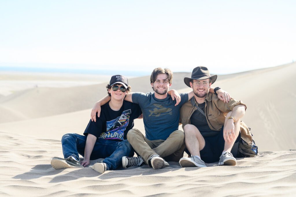 Three young men sitting on the dunes with their arms around each other at the Great Sand Dunes as captured by a Denver Branding photographer and branding strategist.