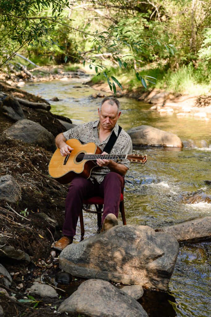 A closeup of a musician sitting on a chair in the middle of a river playing guitar captured by a Denver branding photographer and brand strategist.