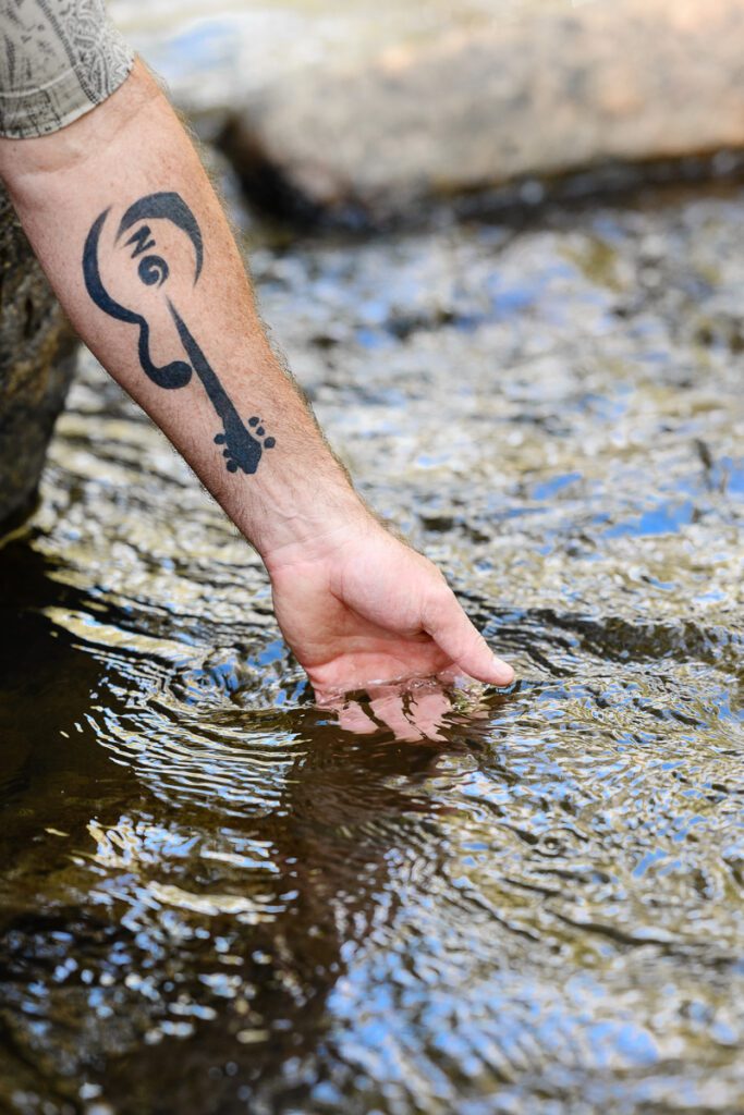 A closeup of a man with a guitar tattoo dipping his hand into a river as captured by a Denver Branding Photographer and brand strategist.