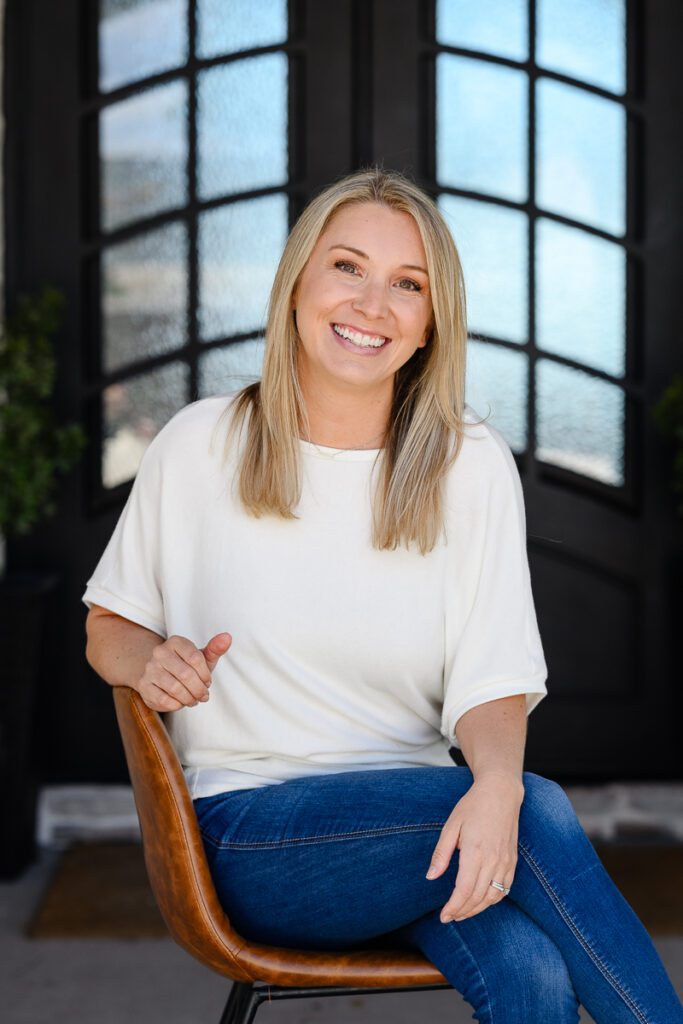 A closeup of a woman in a white shirt sitting on a stool smiling at a Denver Branding Photographer and branding strategist