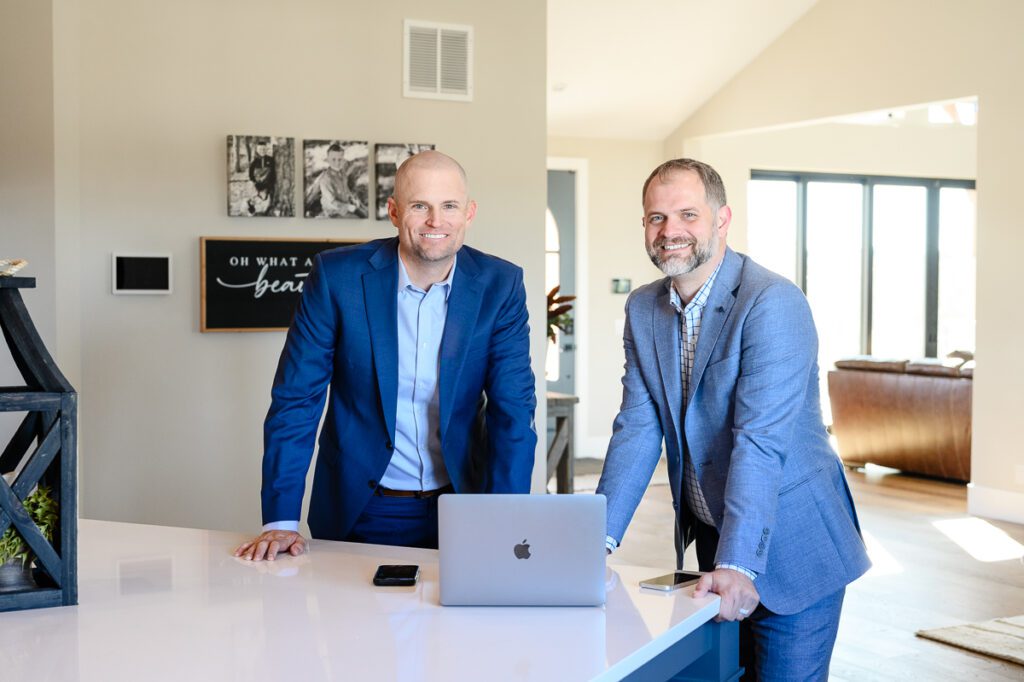 2 men in blue suits standing at a kitchen counter with a laptop smiling at a Denver Branding Photographer for their Denver Branding photos.
