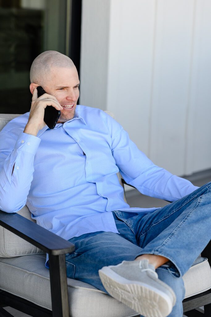 A man in a blue button down shirt sitting in a chair out on a patio smiling while talking on his phone captured by a Denver branding photographer and brand coach.