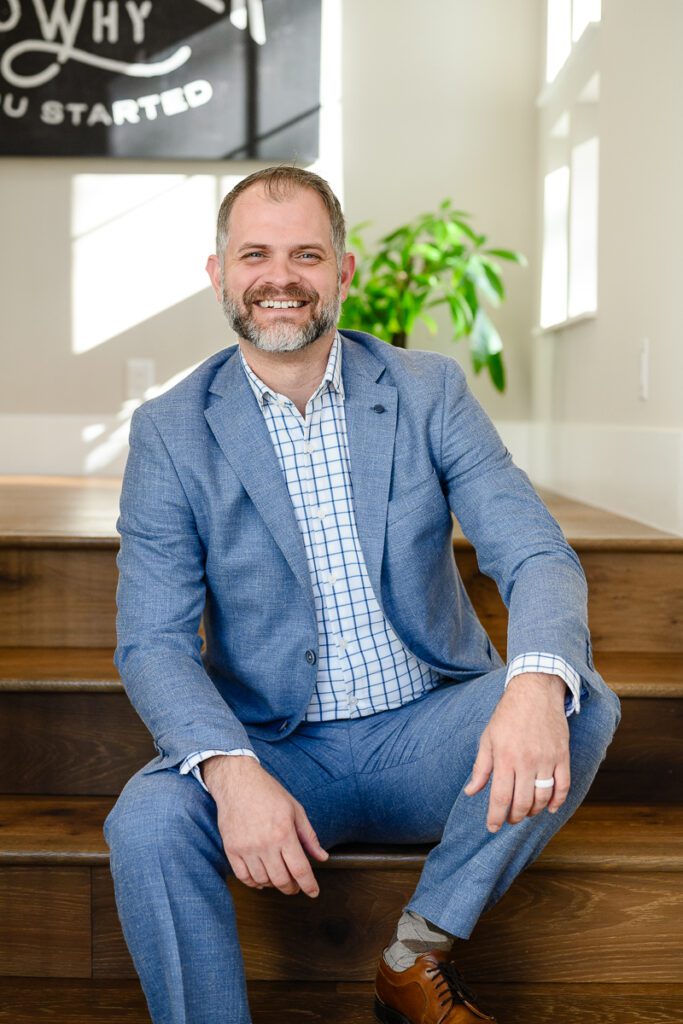 A man in a blue suit sits on the stairs of a home smiling at Denver Branding photographer and branding strategist.