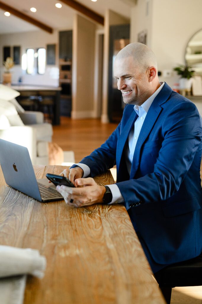 A man in a suit holding a phone smiling at his computer screen captured by Denver branding photographer and branding consultant.