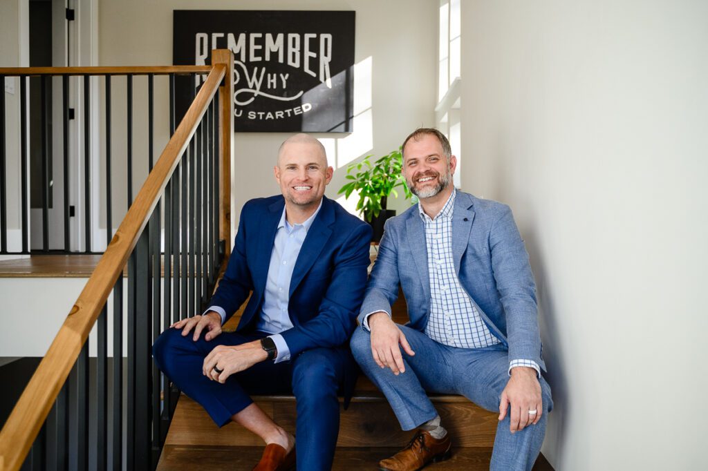 Two men in a blue suit sits on the stairs of a home smiling at Denver Branding photographer and branding strategist.