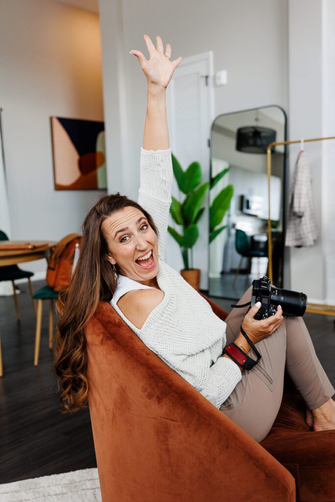A branding consultant, brand coach and brand strategist sits on an orange chair with her feet curled up and and her hand raised in the air with a look of surprise on her face.