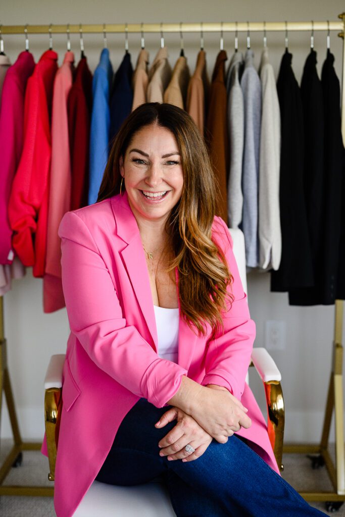 A business owner in a bright pink jack sits in her office surrounded by blazers captured by a Denver branding photographer and Denver commercial photographers.