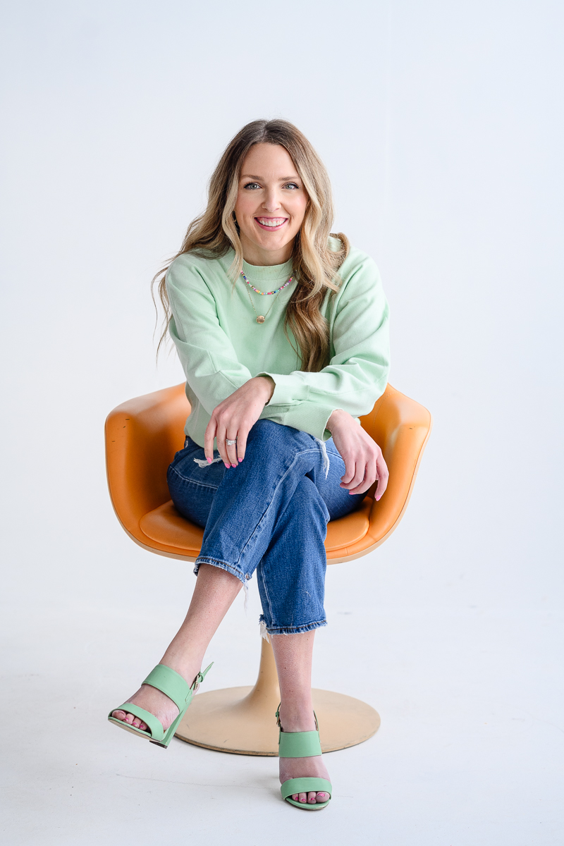small business owner headshot with woman in a mint green sweater sitting on an orange chair and smiling with her legs crossed