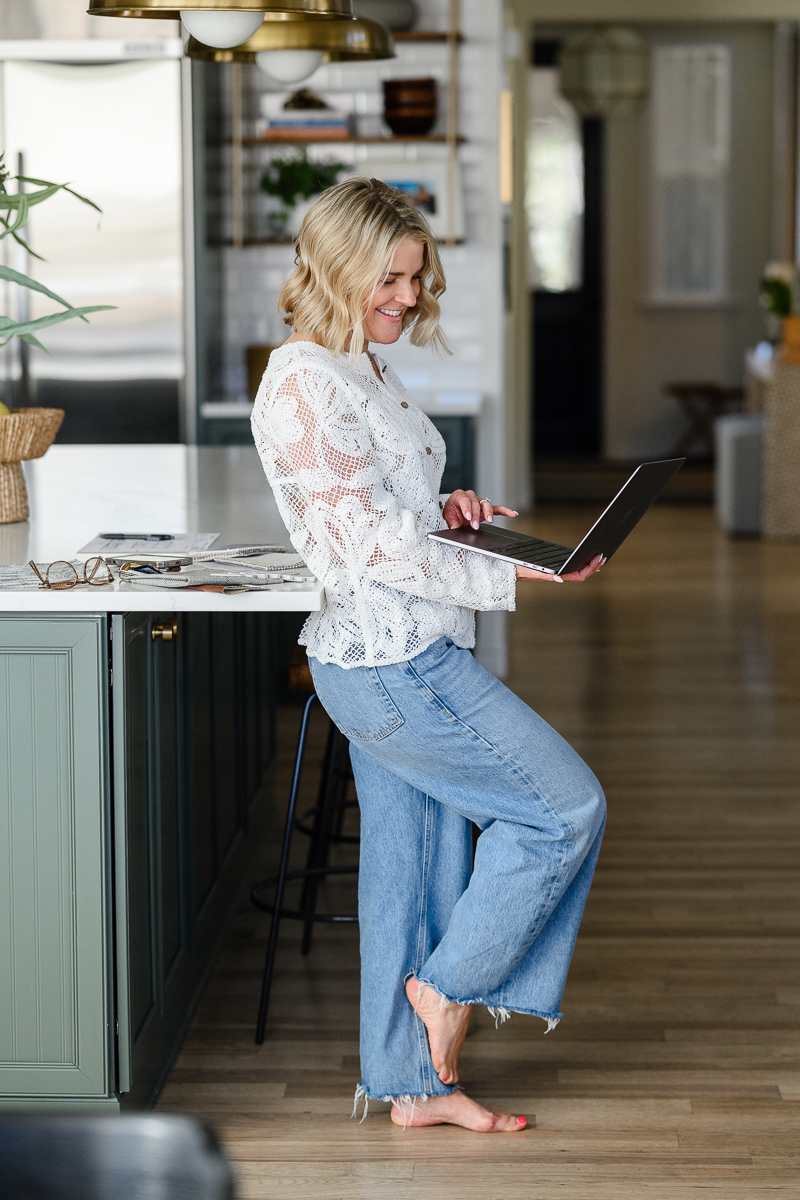 denver commercial photographer captures business woman in a white lace top and blue jeans leaning against a counter in a kitchen and working on her laptop for candid brand photos