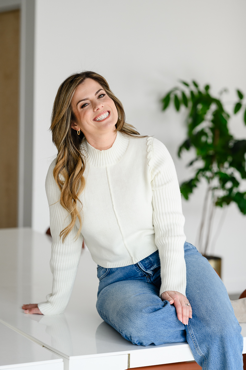 woman in a white sweater and blue jeans sits on a counter in a kitchen and smiles as she leans back while resting her hand on the counter behind her captured by denver commercial photographer