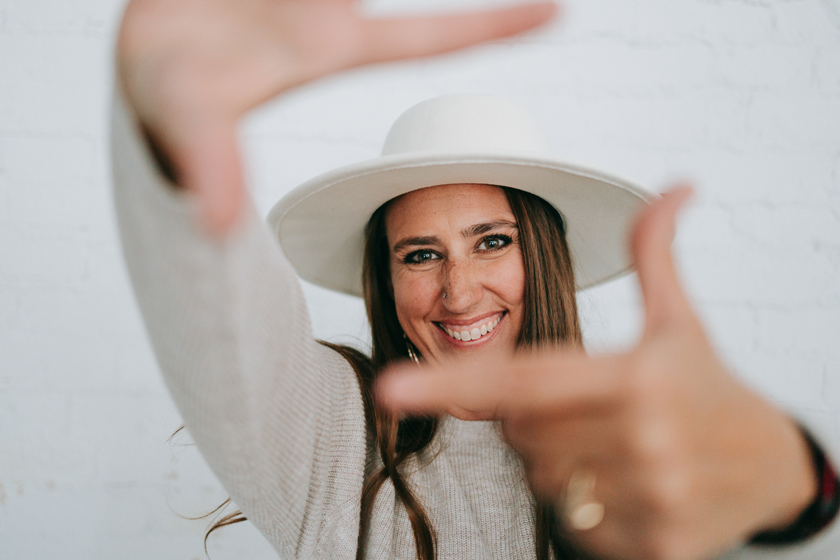 Close-up of a smiling woman wearing a wide-brimmed white hat, playfully framing the camera with her hands, symbolizing creativity and vision in branding photography and the business color meaning of white for clarity and possibility.