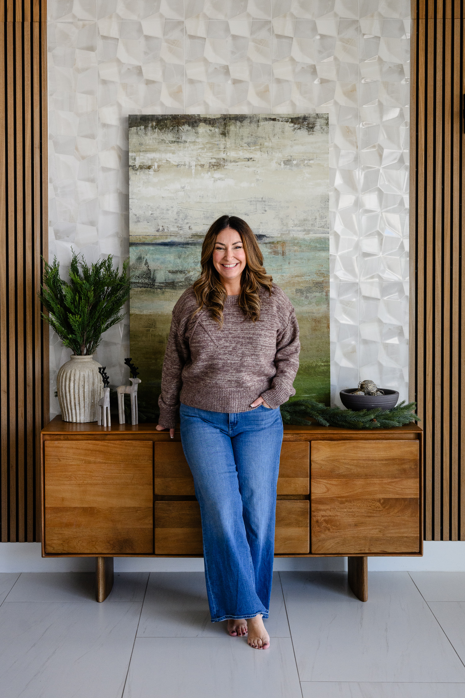 A woman stands barefoot in front of a modern wood console, smiling warmly in a cozy sweater and jeans. The stylish, textured backdrop and subtle seasonal decor make this brand portrait both relaxed and polished—perfectly captured by a Denver brand photographer.